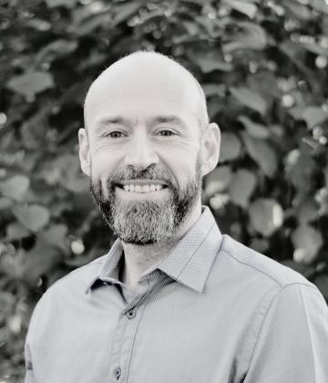 Nick Jones smiling at the camera in a blue shirt, in front of some foliage